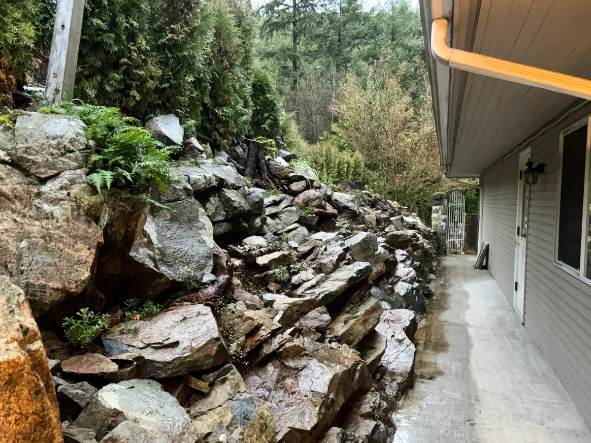 West Vancouver Garden Pond, Paths & Night Lighting — before — Side-of-house rock retaining corridor during construction, then finished lit stone path and planted rock garden along the house wall.