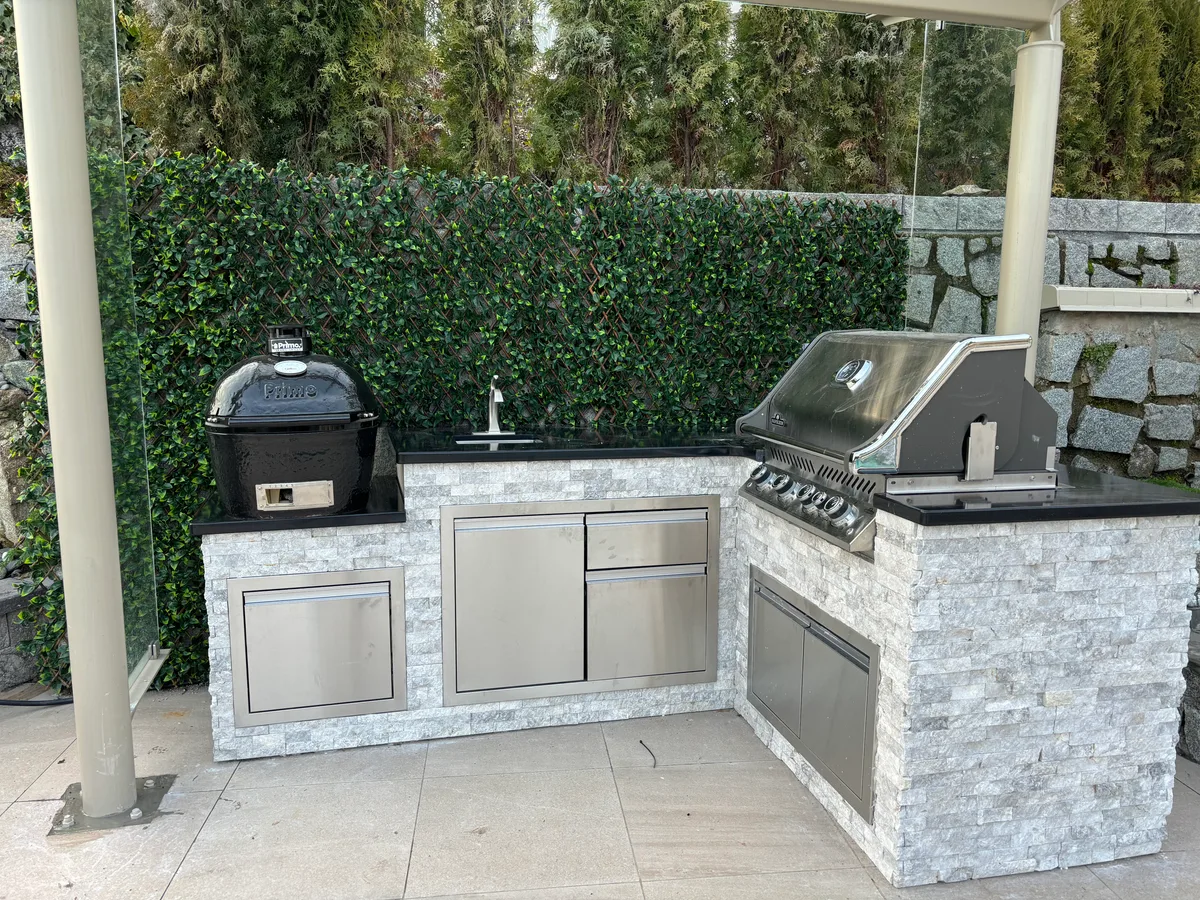 West Vancouver Outdoor Kitchen & Glass Canopy — after — Same stone-wall cooking corner: portable grill and side table, then built-in outdoor kitchen with stainless appliances and stone veneer.