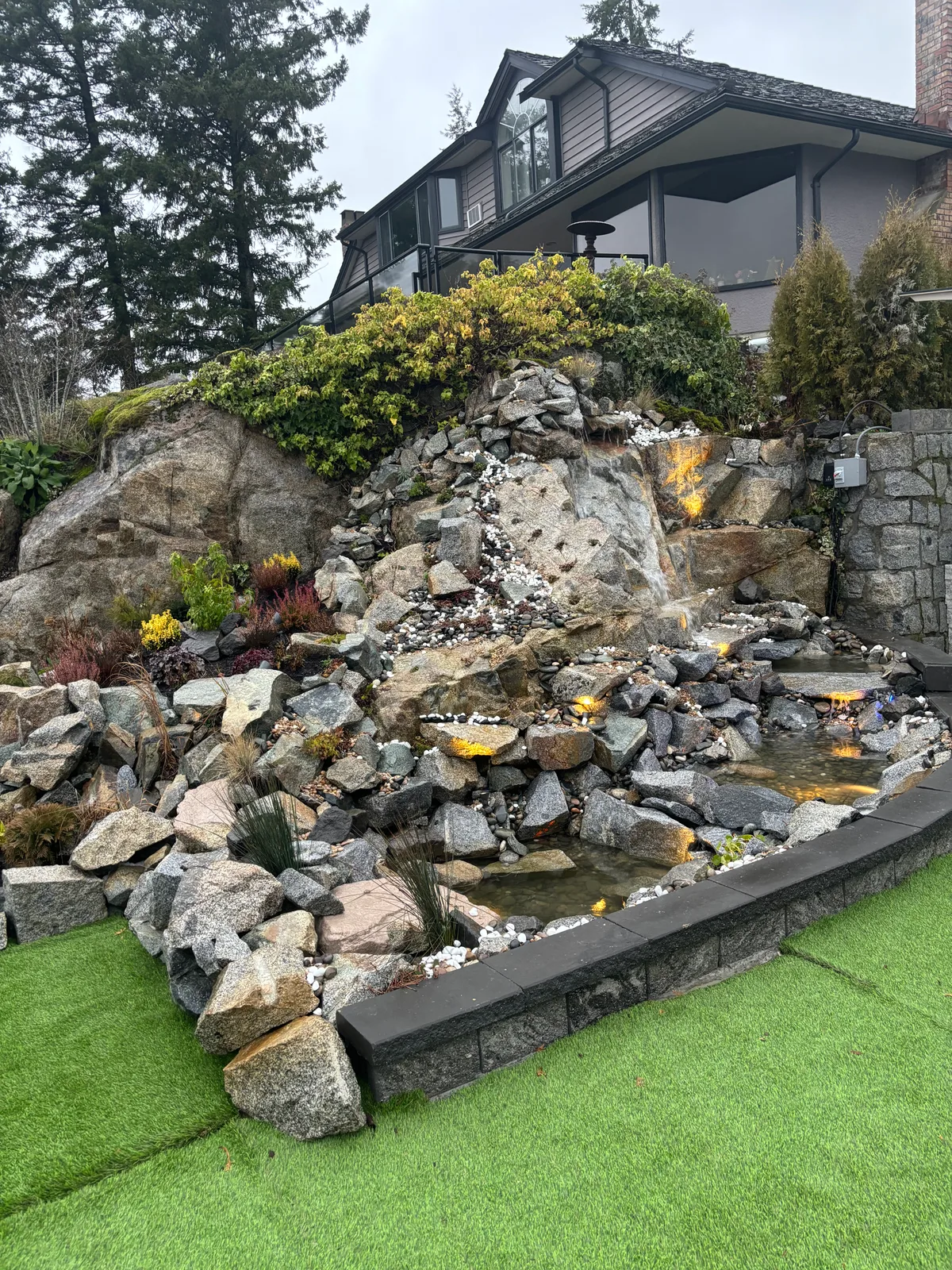 Waterfall under construction beside glass railing and ocean-view trees, then finished flowing rock cascade.