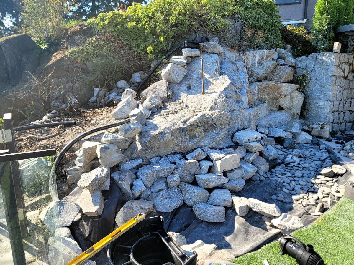 West Vancouver Rock Waterfall & Hillside Hardscape — before — Same waterfall basin during pump and liner install, then completed multi-tier waterfall with turf edge and night lighting.
