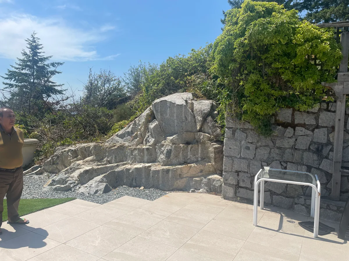 West Vancouver Rock Waterfall & Hillside Hardscape — before — Raw stacked rock face during construction, then completed hillside waterfall with surrounding plantings.